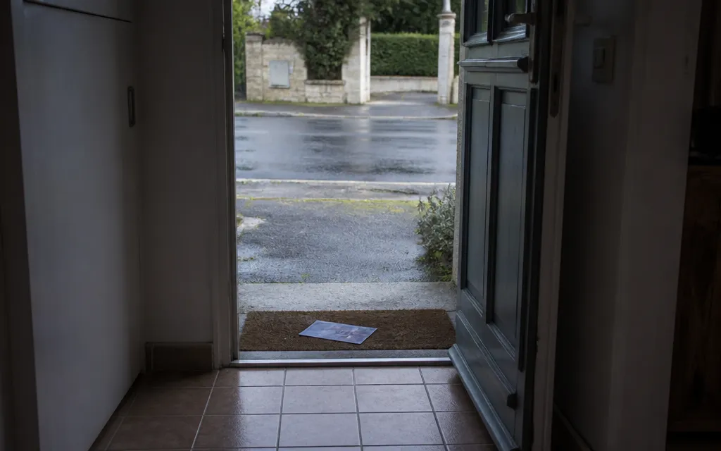 Porte d’entrée entrebâillée sur un pas-de-porte, prospectus glissé dans l’encadrement