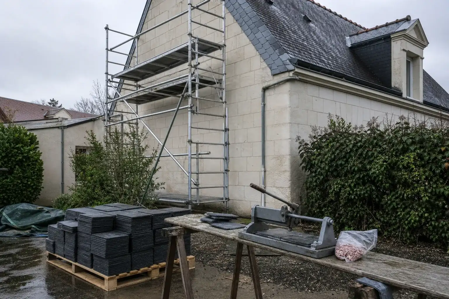 Échafaudage de pied monté contre le pignon d’une maison de faubourg, ardoises en palettes et sac de crochets alignés au sol