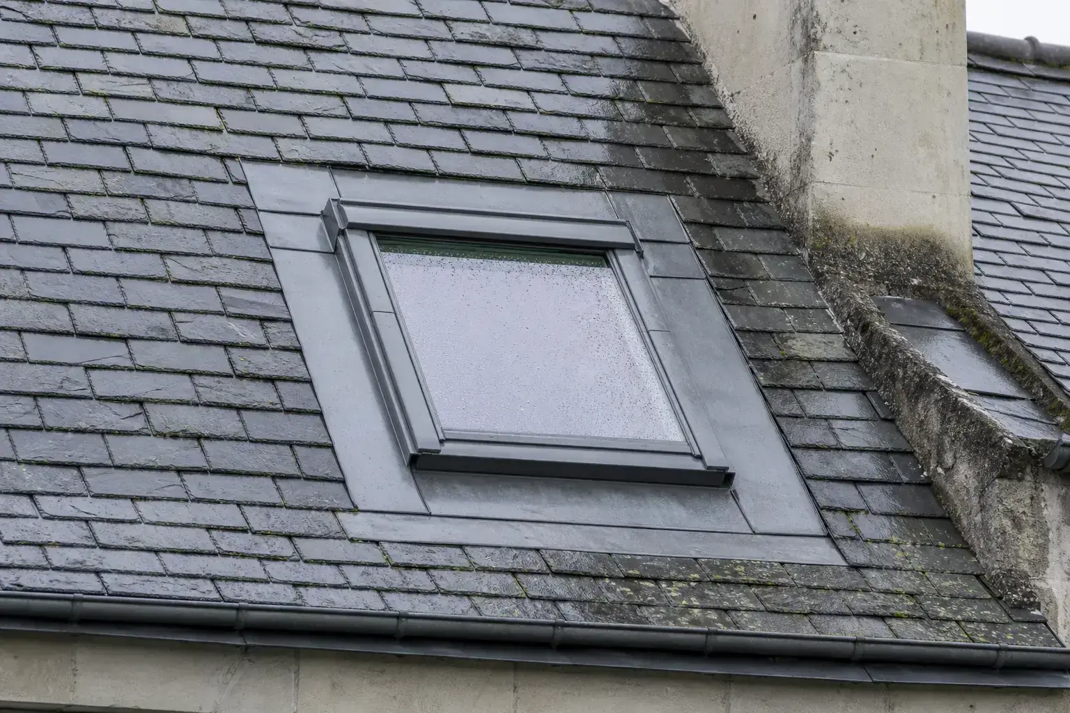 Fenêtre de toit posée dans un versant d’ardoise, bavette de raccord en plomb repliée sur les ardoises du pourtour