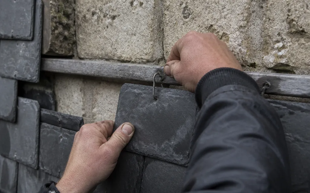Main d’un couvreur enfonçant un crochet inox dans le liteau avant de glisser l’ardoise dessus